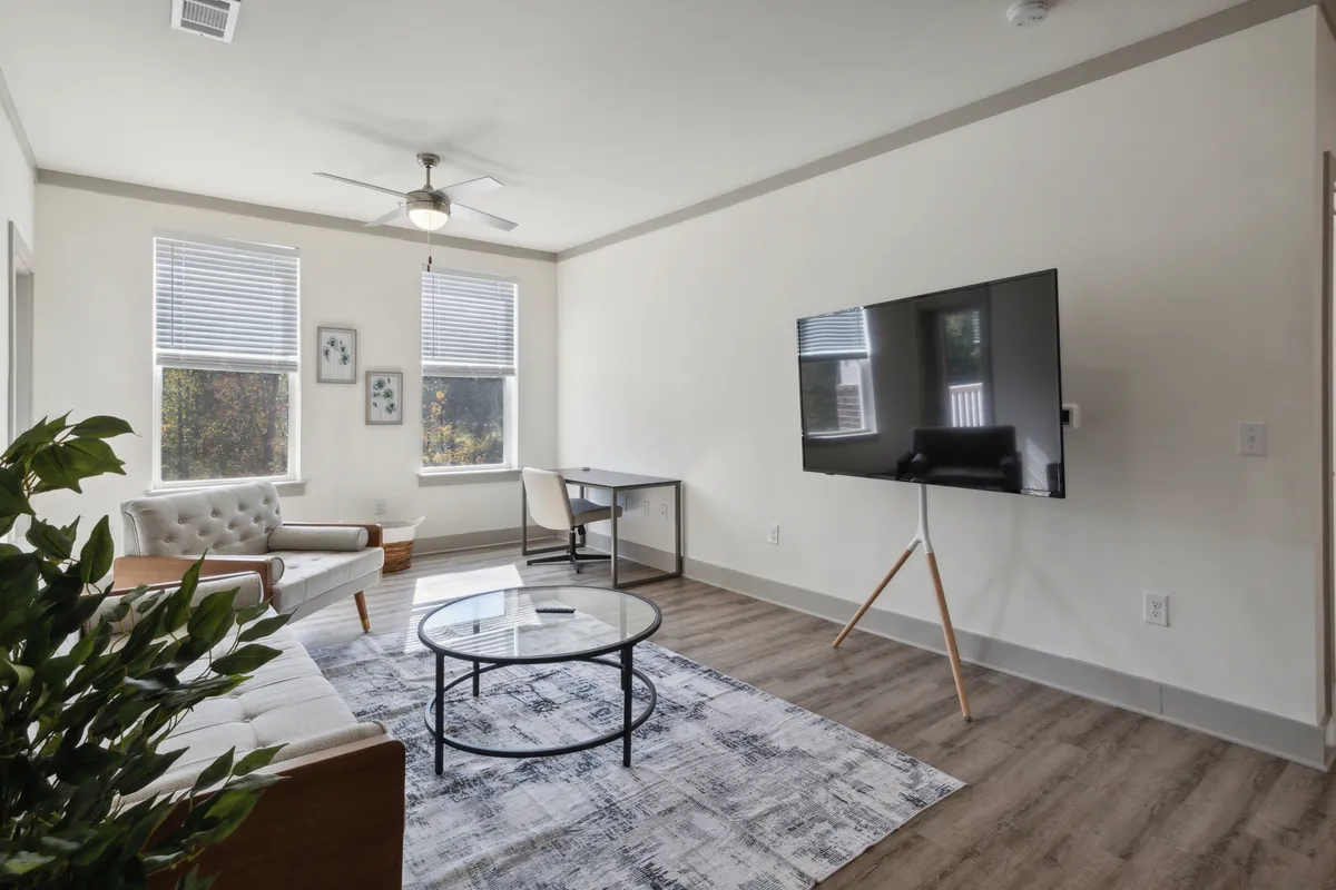 A bright, comfortable living room featuring a plush sofa, modern coffee table, wall-mounted television, and a dedicated desk with a chair. The space is accentuated with soft lighting and neutral tones, creating an inviting atmosphere ideal for both relaxation and productivity.
