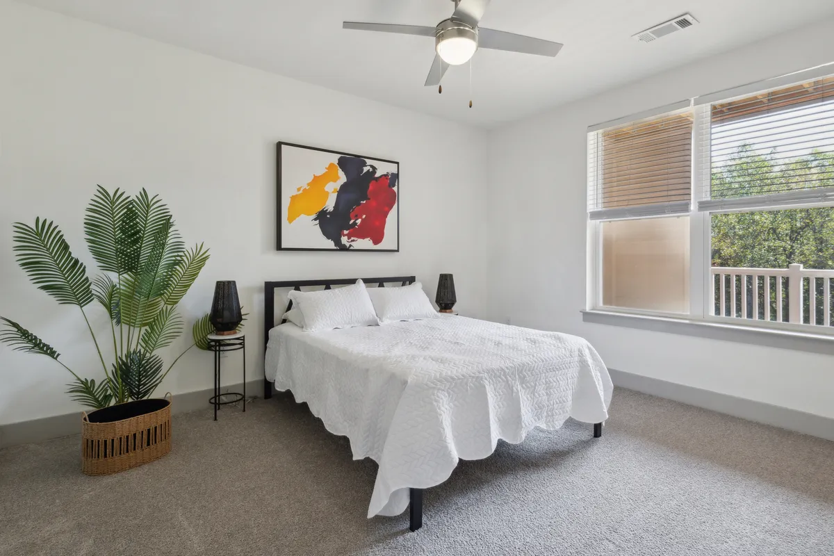 A tranquil bedroom setting with a neatly made bed, tasteful artwork, a green plant, and a soft-glow lamp. The space projects comfort and serenity, suggesting a restful retreat after a busy day.