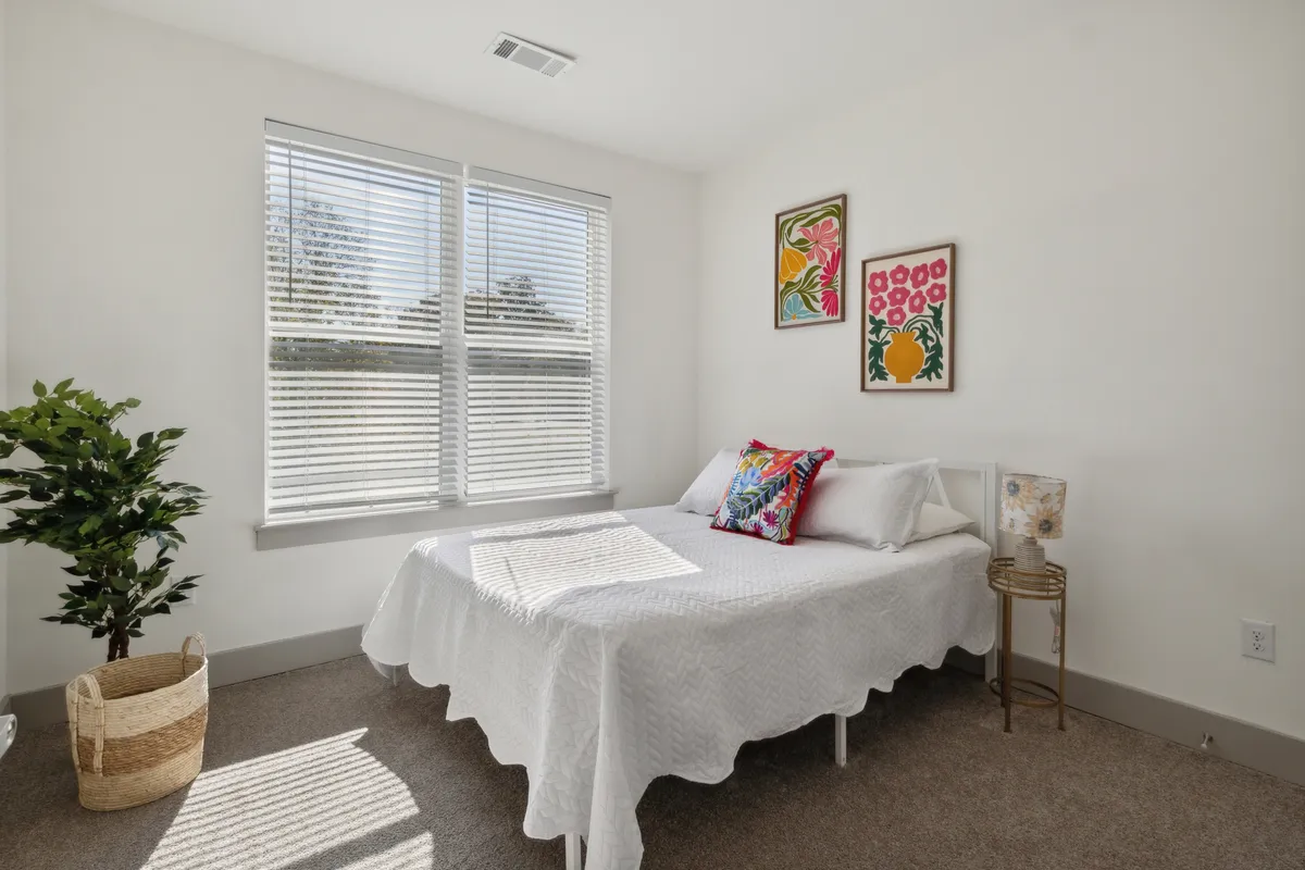 A bright, cozy bedroom featuring a comfortable bed with soft bedding, artwork on the walls, a potted plant for a touch of greenery, and a large window with blinds allowing in natural light. The overall scene suggests a peaceful, restful space ideal for leisurely mornings.