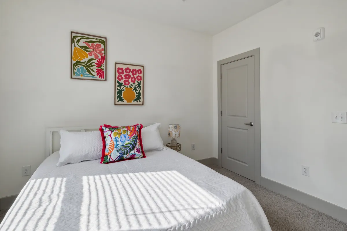 A vibrant bedroom scene with decorative pillows, framed artwork above the bed, and sunlight streaming in. The space feels refreshing and lively while retaining a sense of comfort, making it inviting for relaxation or enjoying a morning coffee.