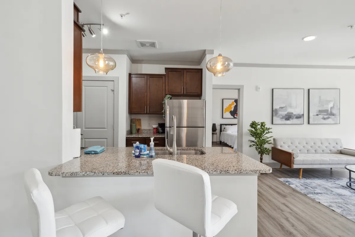 The image offers a view connecting the kitchen and dining area, showcasing a granite countertop, white bar stools, stainless steel appliances, wall artwork, and the adjacent living area. This perspective highlights the integration of dining, cooking, and socializing spaces.